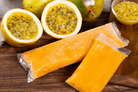 Polpas congeladas de maracujá embaladas ao lado de frutos de maracujá cortados ao meio sobre uma mesa de madeira.