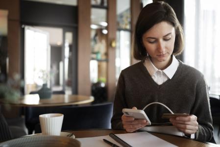 Mulher de camisa branca e suéter marrom usa serviços bancários pelo celular com autenticação em camadas. À sua frente há uma mesa com um copo de café, papéis e canetas.