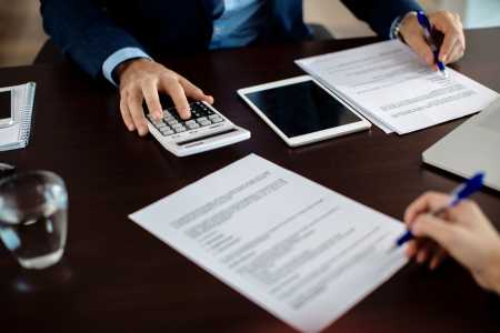 Close-up de duas pessoas analisando documentos sobre uma mesa durante uma reunião. Uma delas utiliza uma calculadora, enquanto a outra aponta informações em um contrato com uma caneta. Sobre a mesa também há um tablet, um notebook, um bloco de notas e um copo com água, sugerindo uma consulta financeira, contábil ou administrativa.