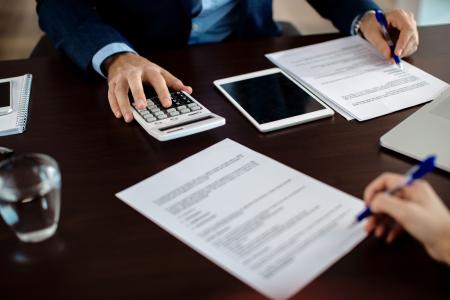 Close-up de duas pessoas analisando documentos sobre uma mesa durante uma reunião. Uma delas utiliza uma calculadora, enquanto a outra aponta informações em um contrato com uma caneta. Sobre a mesa também há um tablet, um notebook, um bloco de notas e um copo com água, sugerindo uma consulta financeira, contábil ou administrativa.