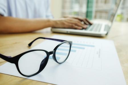 Pessoa usa notebook para realizar transações bancárias, com papel e óculos apoiados em uma mesa.