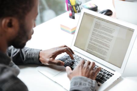 Homem negro utiliza um notebook para ler e editar um documento digital, com as mãos sobre o teclado, em uma mesa de trabalho com materiais de escritório ao fundo.