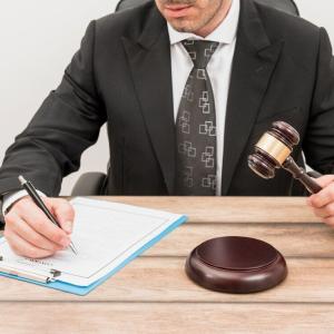Homem de terno preto e gravata estampada sentado à mesa enquanto preenche um documento em uma prancheta com uma caneta. Na outra mão, segura um martelo de juiz acima de uma base de madeira, simbolizando decisões judiciais ou processos legais.