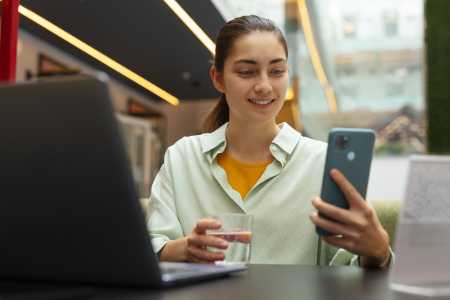 Mulher de camisa branca sorri com copo de água transparente e celular nas mãos, com cotovelos apoiados em mesa à frente dela, com notebook em cima.