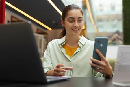 Mulher de camisa branca sorri com copo de água transparente e celular nas mãos, com cotovelos apoiados em mesa à frente dela, com notebook em cima.