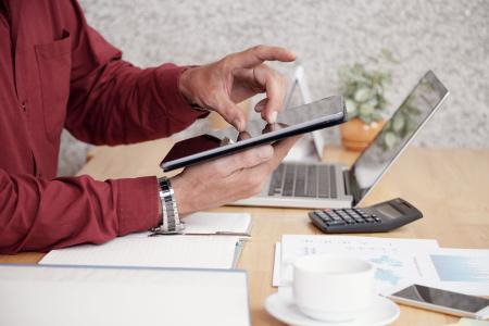 Pessoa usa tablet para utilizar serviços bancários digitais. Há uma mesa com notebook, calculadora, um vaso de plantas, uma xícara de café e papéis ao lado da pessoa.