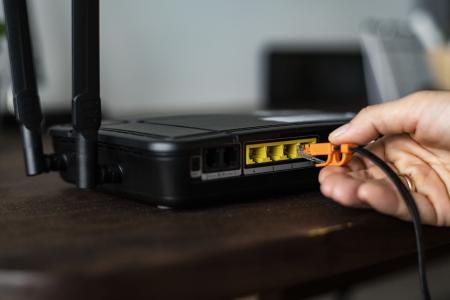 Close-up de uma mão conectando um cabo Ethernet laranja a uma das portas LAN de um roteador Wi-Fi preto com duas antenas. O roteador está sobre uma mesa de madeira, com as demais portas de rede visíveis ao lado.