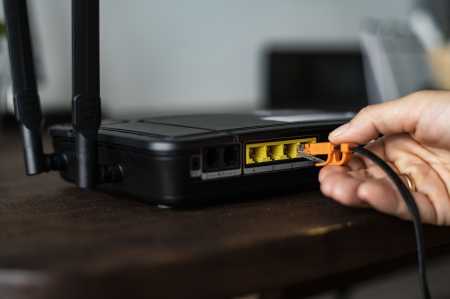 Close-up de uma mão conectando um cabo Ethernet laranja a uma das portas LAN de um roteador Wi-Fi preto com duas antenas. O roteador está sobre uma mesa de madeira, com as demais portas de rede visíveis ao lado.