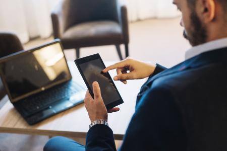 Homem sentado em uma mesa utilizando um tablet enquanto um notebook permanece aberto ao lado. A imagem destaca o uso de dispositivos digitais em um ambiente de trabalho ou escritório, sugerindo atividades como gestão de documentos, consultas online ou serviços digitais.