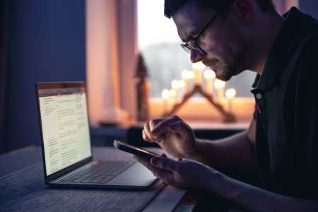 Homem de óculos utiliza um smartphone enquanto consulta informações em um notebook sobre a mesa, em um ambiente interno durante a noite, iluminado pela luz da tela e por velas ao fundo.