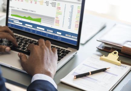 Pessoa usa serviços financeiros pelo notebook, que está apoiado em cima de uma mesa, com papéis e caneta ao lado.