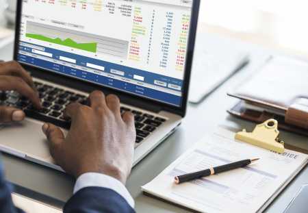 Pessoa usa serviços financeiros pelo notebook, que está apoiado em cima de uma mesa, com papéis e caneta ao lado.