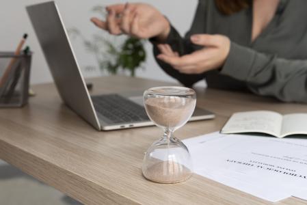 Ampulheta sobre uma mesa de escritório, ao lado de um notebook e documentos, enquanto uma pessoa desfocada gesticula durante uma conversa. A imagem representa a importância dos prazos, da gestão do tempo e da organização de processos em um ambiente profissional.