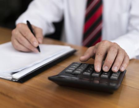 Homem utiliza uma calculadora enquanto faz anotações em um documento sobre uma mesa, representando o cálculo de despesas, orçamento ou planejamento financeiro.
