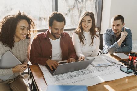 Grupo de quatro pessoas reunidas em uma mesa de trabalho analisando informações em um notebook. Ao redor, há documentos, plantas e materiais de escritório, representando colaboração, planejamento e tomada de decisões em equipe em um ambiente corporativo.
