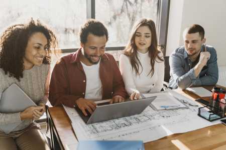 Grupo de quatro pessoas reunidas em uma mesa de trabalho analisando informações em um notebook. Ao redor, há documentos, plantas e materiais de escritório, representando colaboração, planejamento e tomada de decisões em equipe em um ambiente corporativo.