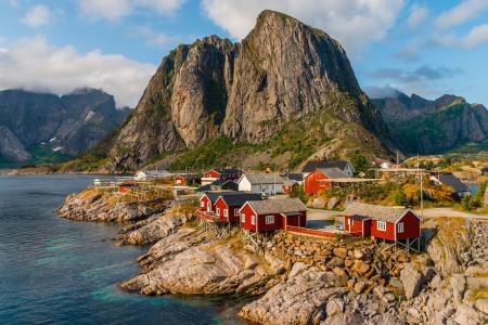 A imagem retrata uma vila costeira cercada por montanhas imponentes, com pequenas casas de madeira vermelhas construídas sobre rochas às margens de um mar de águas calmas e cristalinas. Ao fundo, os grandes paredões rochosos dominam a paisagem, enquanto o céu parcialmente nublado e a vegetação esparsa completam o cenário natural. A cena transmite tranquilidade e destaca a integração entre a arquitetura tradicional e a natureza, sendo uma fotografia típica de regiões costeiras do norte da Europa e frequentemente utilizada para ilustrar temas relacionados a turismo, paisagens naturais e qualidade de vida.