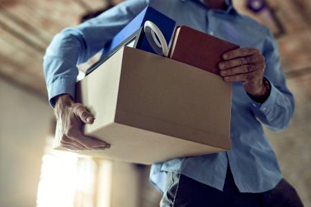 Pessoa carrega uma caixa com pertences pessoais, como pastas e caderno, ao deixar um ambiente de trabalho.