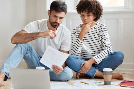 Casal sentado no chão analisa documentos financeiros enquanto consulta um notebook. O homem segura uma folha e uma caneta, enquanto a mulher observa atentamente. Há papéis, um lápis e copos de café ao redor, sugerindo planejamento financeiro ou organização de despesas domésticas.