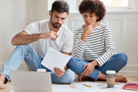 Casal sentado no chão analisa documentos financeiros enquanto consulta um notebook. O homem segura uma folha e uma caneta, enquanto a mulher observa atentamente. Há papéis, um lápis e copos de café ao redor, sugerindo planejamento financeiro ou organização de despesas domésticas.