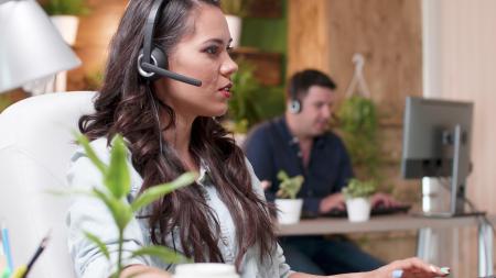Mulher usando headset atende a uma chamada enquanto trabalha em frente ao computador em um escritório. Ao fundo, outro profissional também utiliza um headset diante de um monitor, em um ambiente moderno com plantas e iluminação natural, sugerindo uma central de atendimento ou equipe de suporte ao cliente.
