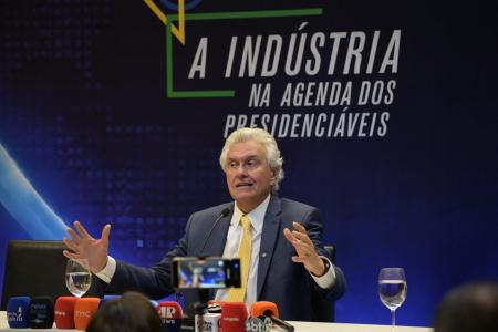 De terno azul escuro, gravata amarela e camisa branca, Ronaldo Caiado fala em microfone sentado em mesa com microfones de diversas emissoras