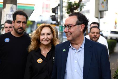 De óculos, blazer azul escuro e camisa social azul clara, Augusto Cury figura de perfil em foto com pessoas da sua campanha ao fundo