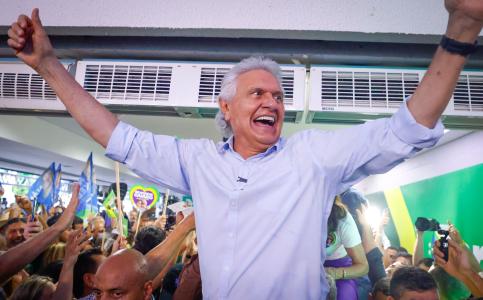 Ronaldo Caiado, de blusa social azul clara, de braços abertos sendo erguido por populares