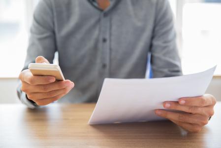 Uma pessoa analisa um documento enquanto utiliza o celular, em um ambiente corporativo, representando uma situação de consulta, conferência ou organização de informações e documentos.