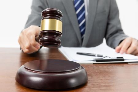 A imagem mostra um juiz ou leiloeiro em um ambiente formal, segurando um martelo de madeira sobre a mesa, com documentos e uma caneta ao fundo, remetendo a processos judiciais, leilões, decisões legais e negociações de bens.