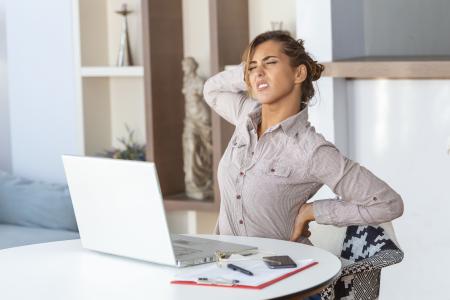 Retrato de uma jovem estressada sentada à mesa do escritório em casa, em frente ao laptop, tocando as costas doloridas com uma expressão de dor, sofrendo de cefaleia após trabalhar no computador.