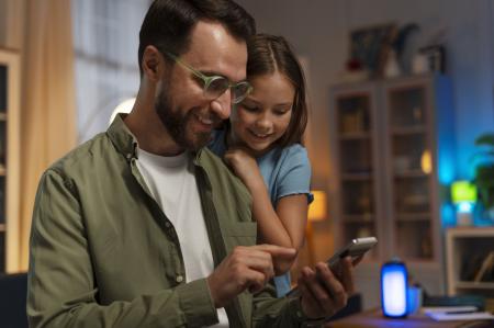 Um pai e uma criança sorriem enquanto olham para um celular, em um ambiente interno aconchegante, sugerindo um momento de convivência e interação digital em família. O homem usa óculos, camiseta branca e camisa verde aberta, enquanto a criança, apoiada em seus ombros, observa a tela com expressão de curiosidade. Ao fundo, há uma estante, iluminação suave e um dispositivo inteligente aceso, reforçando a atmosfera doméstica e tecnológica.