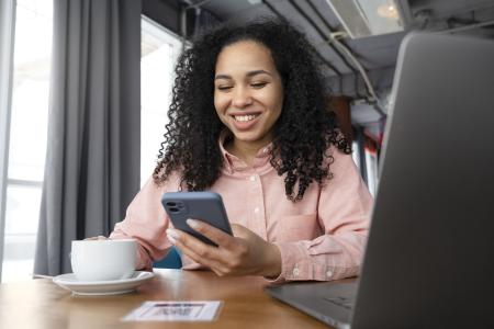 Uma mulher sorri enquanto utiliza o celular em um ambiente de trabalho ou cafeteria, com um notebook aberto e uma xícara de café sobre a mesa. A cena remete ao uso de serviços digitais, compras e pagamentos online, aplicativos financeiros e comunicação, sendo frequentemente utilizada para ilustrar conteúdos sobre tecnologia, finanças e praticidade no dia a dia.