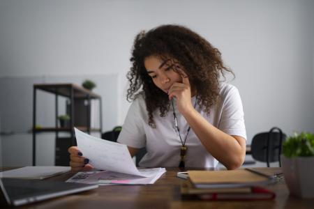 Uma pessoa analisa atentamente documentos sobre uma mesa de trabalho, em um ambiente de escritório ou home office.
