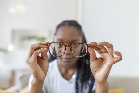 Uma jovem africana segura óculos com lentes de dioptria e olha através deles, representando o problema da miopia e a necessidade de correção visual.