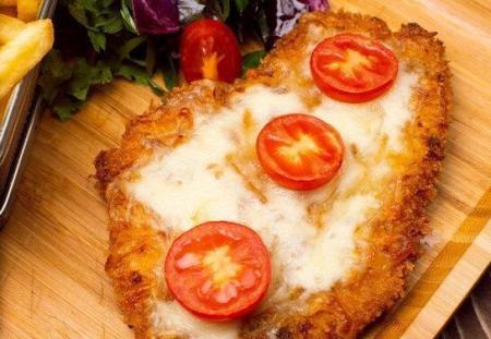 Um filé empanado coberto com queijo derretido e rodelas de tomate é servido em uma tábua de madeira, acompanhado de batatas fritas e uma salada de folhas verdes.