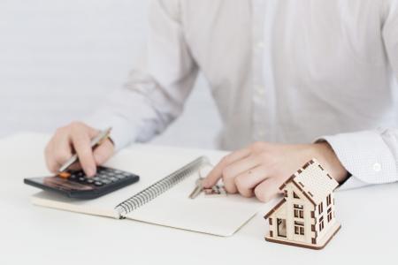 Uma pessoa realiza cálculos relacionados à compra ou negociação de um imóvel, utilizando uma calculadora e documentos de apoio, enquanto uma miniatura de uma casa reforça o contexto imobiliário.