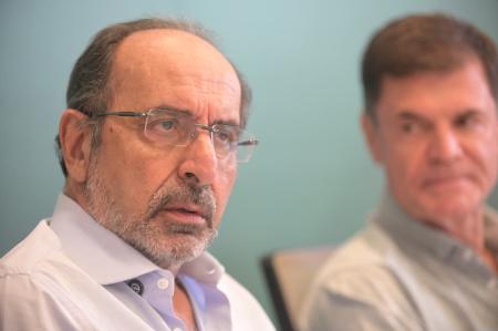 Foto de Alexandre Kalil em primeiro plano (à esq.) com homem branco desfocado à direita, ambos à frente de fundo verde