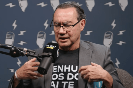 De blazer cinza e camisa preta, com frase estampada, fala em microfone do Flow News