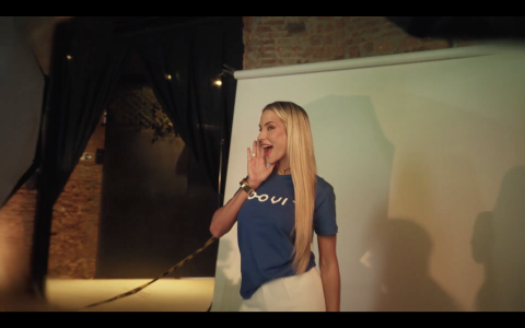 Fotografia mostra gravação de propaganda da Loovi. Centralizada, uma mulher branca de cabelos longos e loiros usa camiseta azul com a marca Loovi. Atrás dela, painel branco