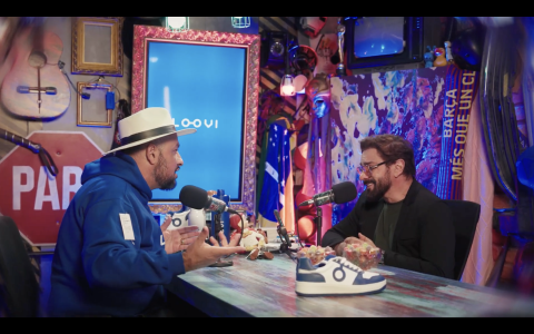 Em cenário de podcast, dois homens se posicionam um à frente do outro em conversa. À esquerda, uma homem negro usando chapéu e moletom azul. À direita, um homem branco de óculos e barba usa camiseta e paletó pretos