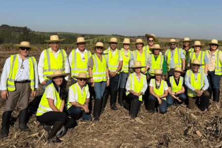 Grupo de pessoas usando coletes de segurança e chapéus posa para foto em uma área rural, com lavoura e vegetação ao fundo.