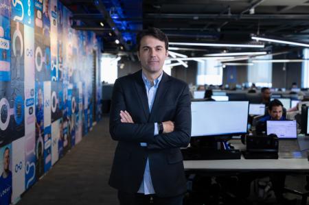 Quezide Cunha, CEO da Loovi, sorrindo e de braços cruzados no escritório corporativo da Loovi, com computadores ao fundo.