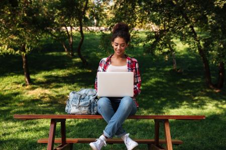 Estudante sentada em banco em ambiente com paisagem verde enquanto utiliza serviços financeiros globais pelo notebook.