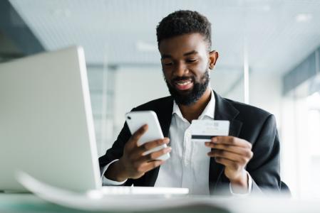 Homem de camisa branca e blazer com celular e cartão de banco nas mãos.