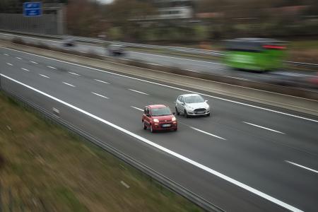 Imagem mostra a autoestrada com carros passando desfocados em velocidade. Em foco, dois carros, lado a lado, seguindo a mesma direção. O carro à esquerda é vermelho e o da direita é branco