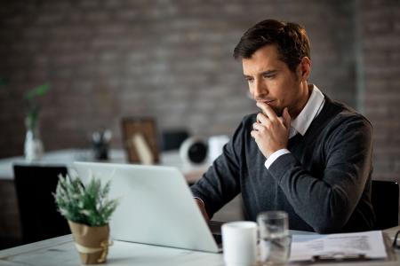 Um homem com camisa social branca e suéter cinza está sentado pensativo em uma mesa rústica, com a mão no queixo e os olhos fixos na tela de um notebook aberto à sua frente. Ao redor dele, vê-se um ambiente de escritório com uma parede de tijolos ao fundo, uma plantinha em um vaso, uma xícara, um copo de água e documentos, todos em foco ou ligeiramente desfocados.