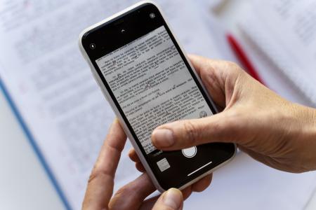 Mãos seguram um smartphone que exibe um texto, sobre uma mesa com documentos e uma caneta vermelha ao fundo.