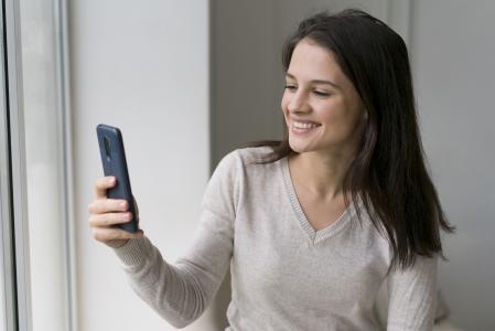 Mulher sorrindo enquanto segura um smartphone e desbloqueia o celular por reconhecimento facial próxima a uma janela.