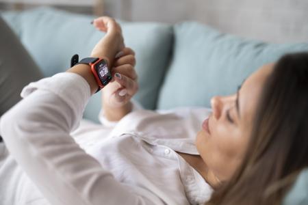 Mulher deitada em um sofá usando um smartwatch no pulso, com a tela do dispositivo exibindo o horário.